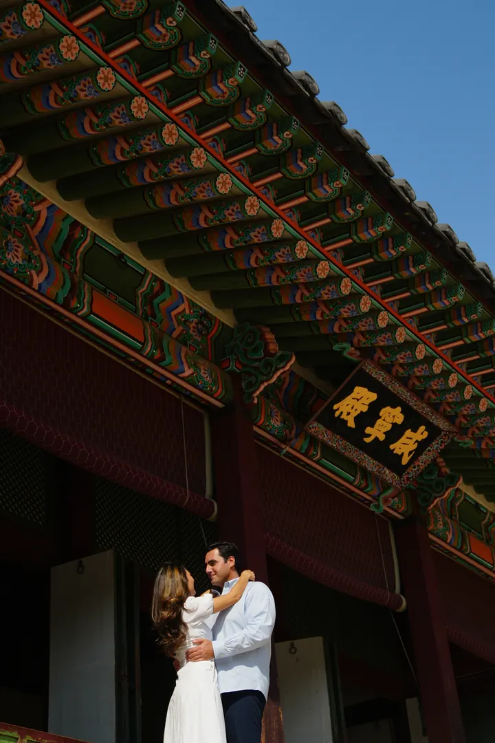 Connor and Diane embrace beneath the painted eaves of a Korean palace hall against a clear blue sky.